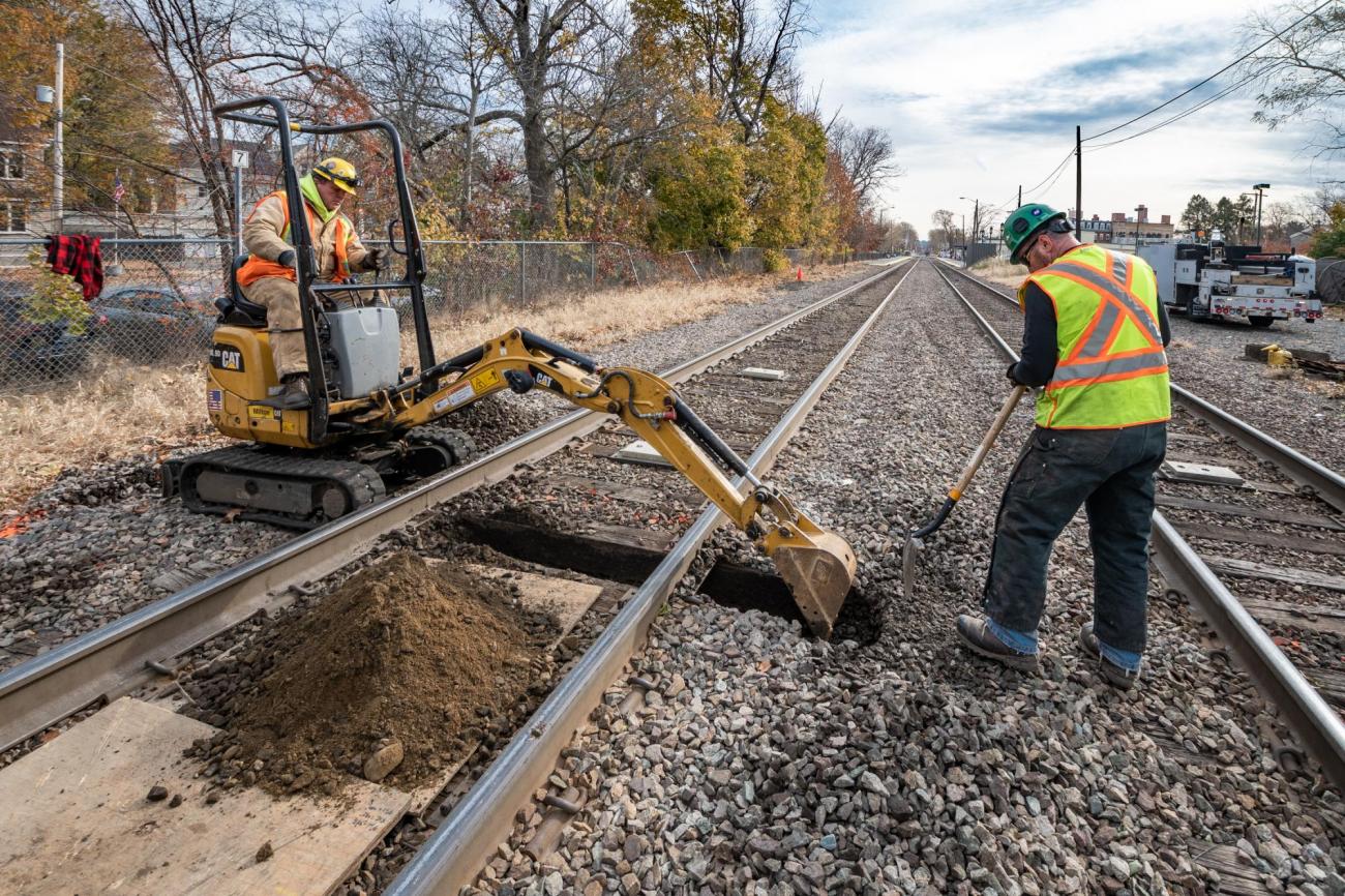 Crew members excavate under the track to prepare for power line installation as part of the PTC upgrades on the Lowell Line of the Commuter Rail (November 2019)