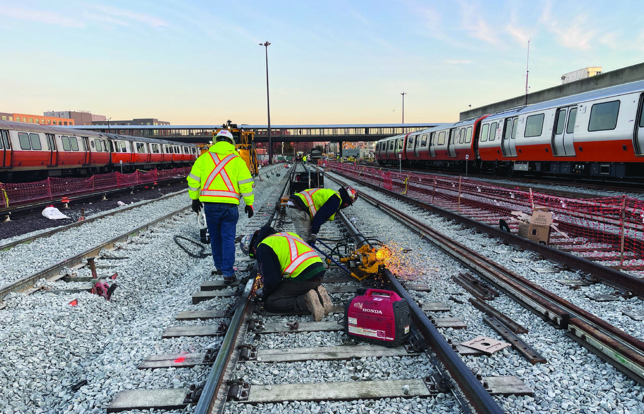 3 crew members welding tracks. there are orange line trains on other tracks to the left and right of the tracks they're working on