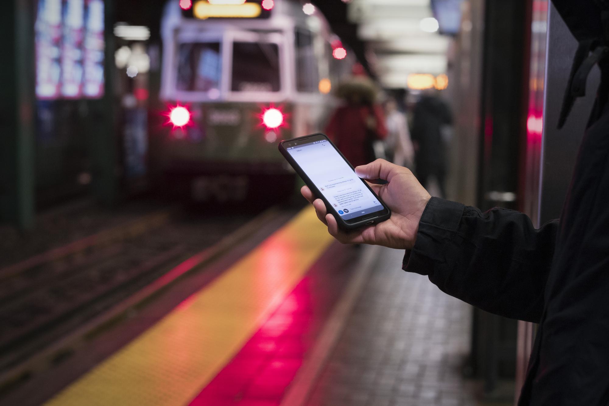 On the Green Line platform at Park Street, a rider holds a smartphone, looking at a T-Alerts text message.
