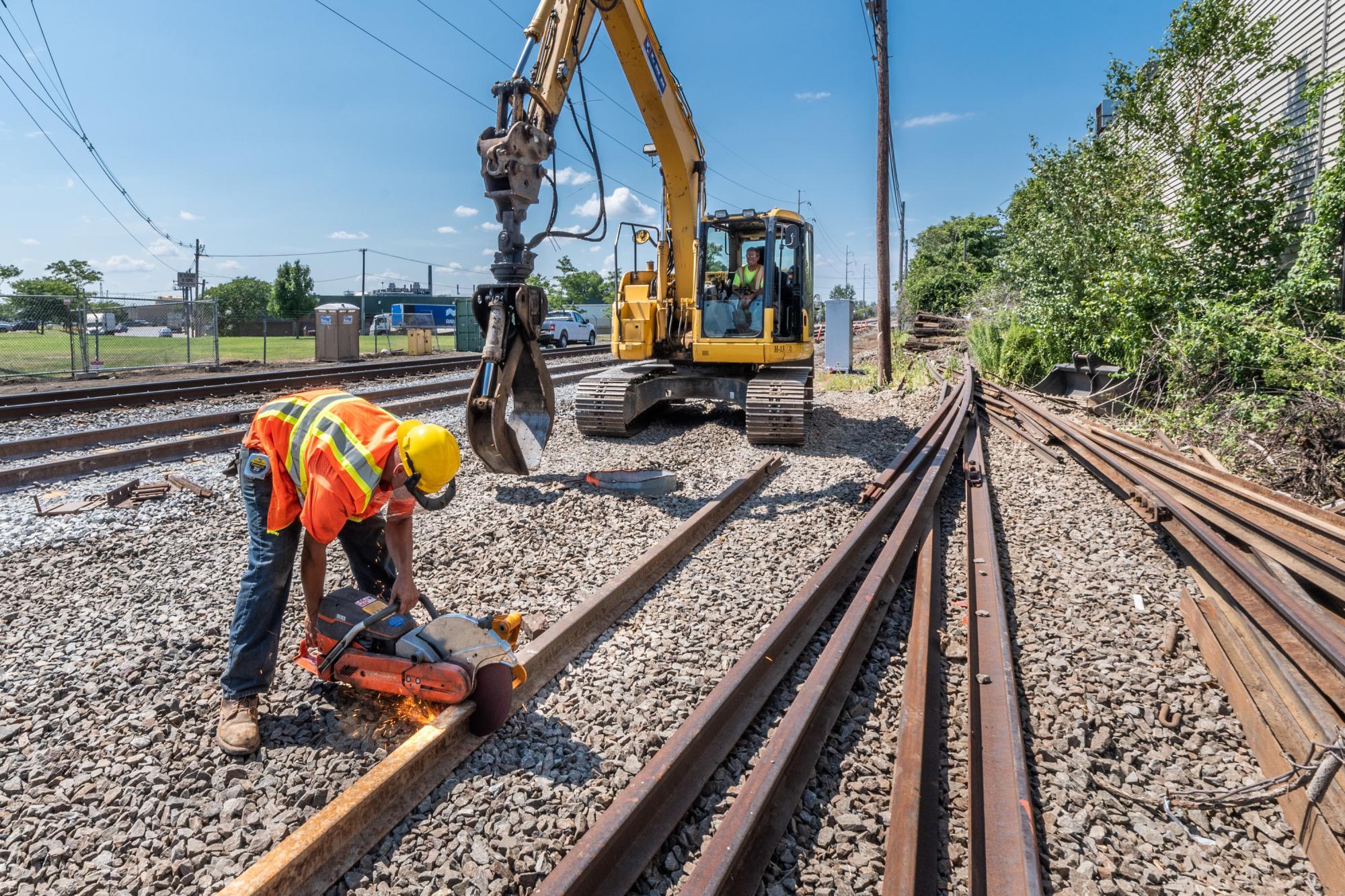 A crewperson uses a handheld rail cutting machine to cut steel rail, with an excavator in the background, and lengths of rail on the right.