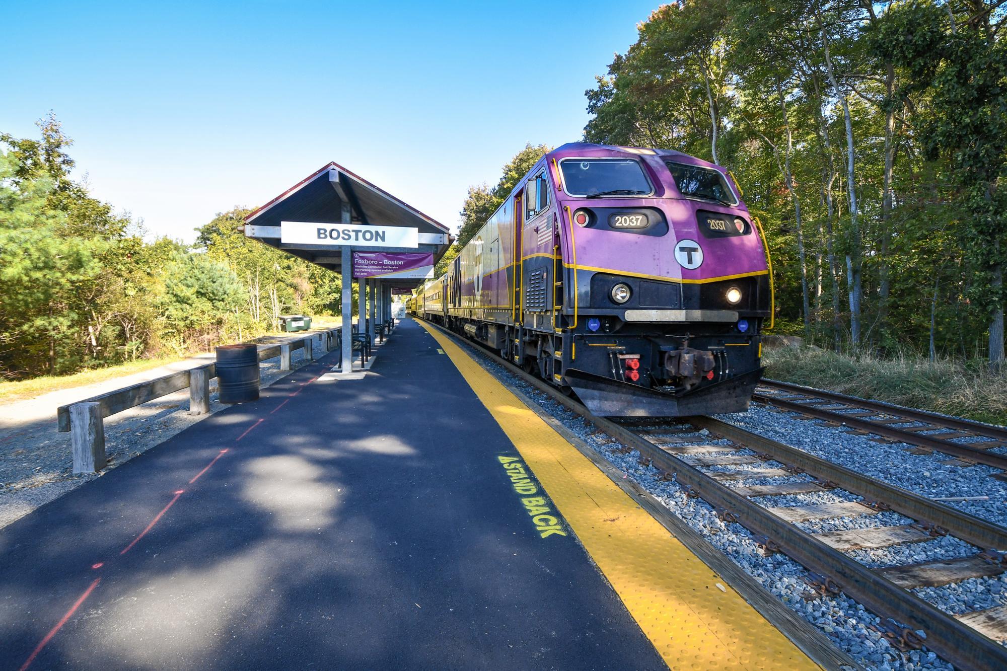 A Commuter Rail train pulls away from Foxboro Station, inbound for Boston. In the background is a sign adveristing the Foxboro - Boston weekday pilot.
