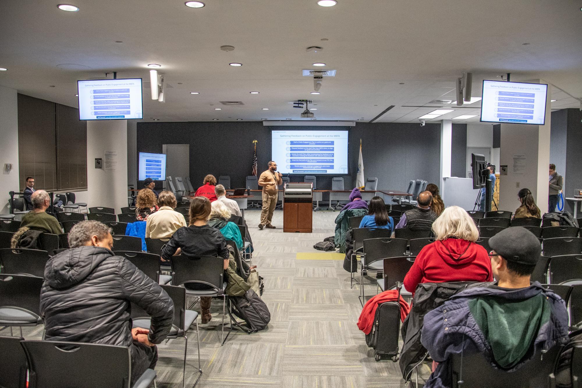 Community members at a public meeting at the State Transportation Building, with 3 screens showing meeting presentation slides and a speaker at the front.
