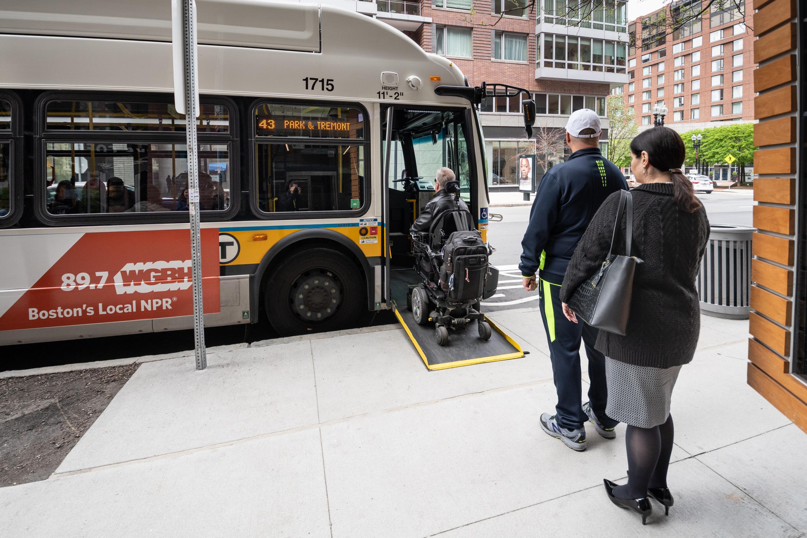 At a bus stop, a rider in a wheeleed mobility device boards the bus via a ramp, as two other riders wait in line behind.