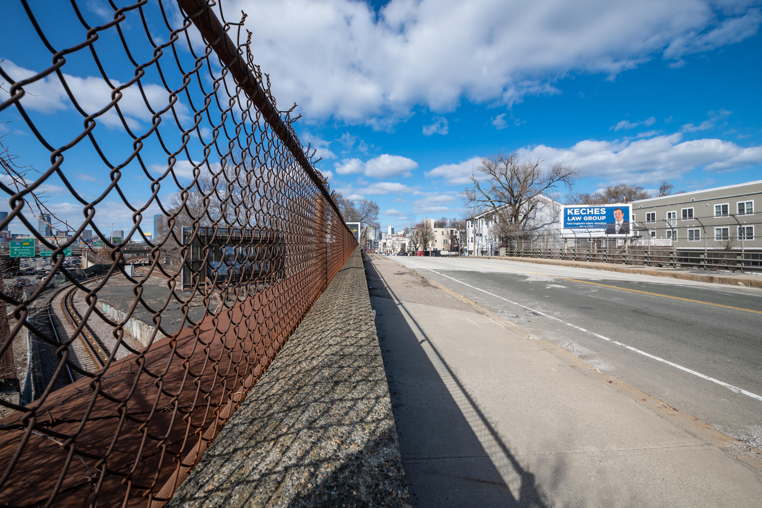 A photo taken on top of the Dorchester Avenue bridge. A highway and train tracks are visible to the left, through a chain link fence.