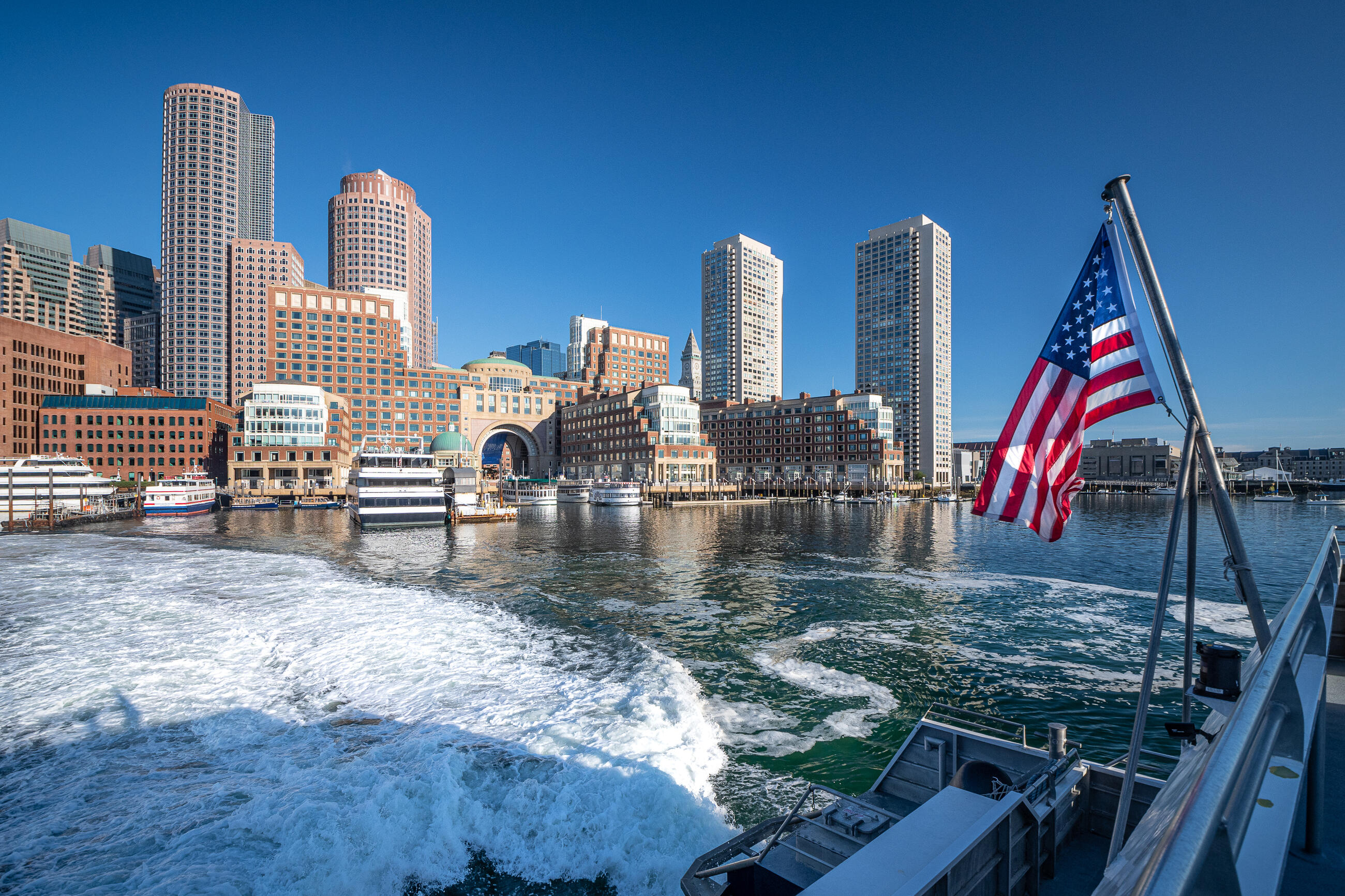 A photo of Rowes Wharf, taken from the ferry across the water. The water is white from the wake of the ferry moving in the water. A United States flag is flying at an angle from the side of the boat, and the sky is a bright clear blue 