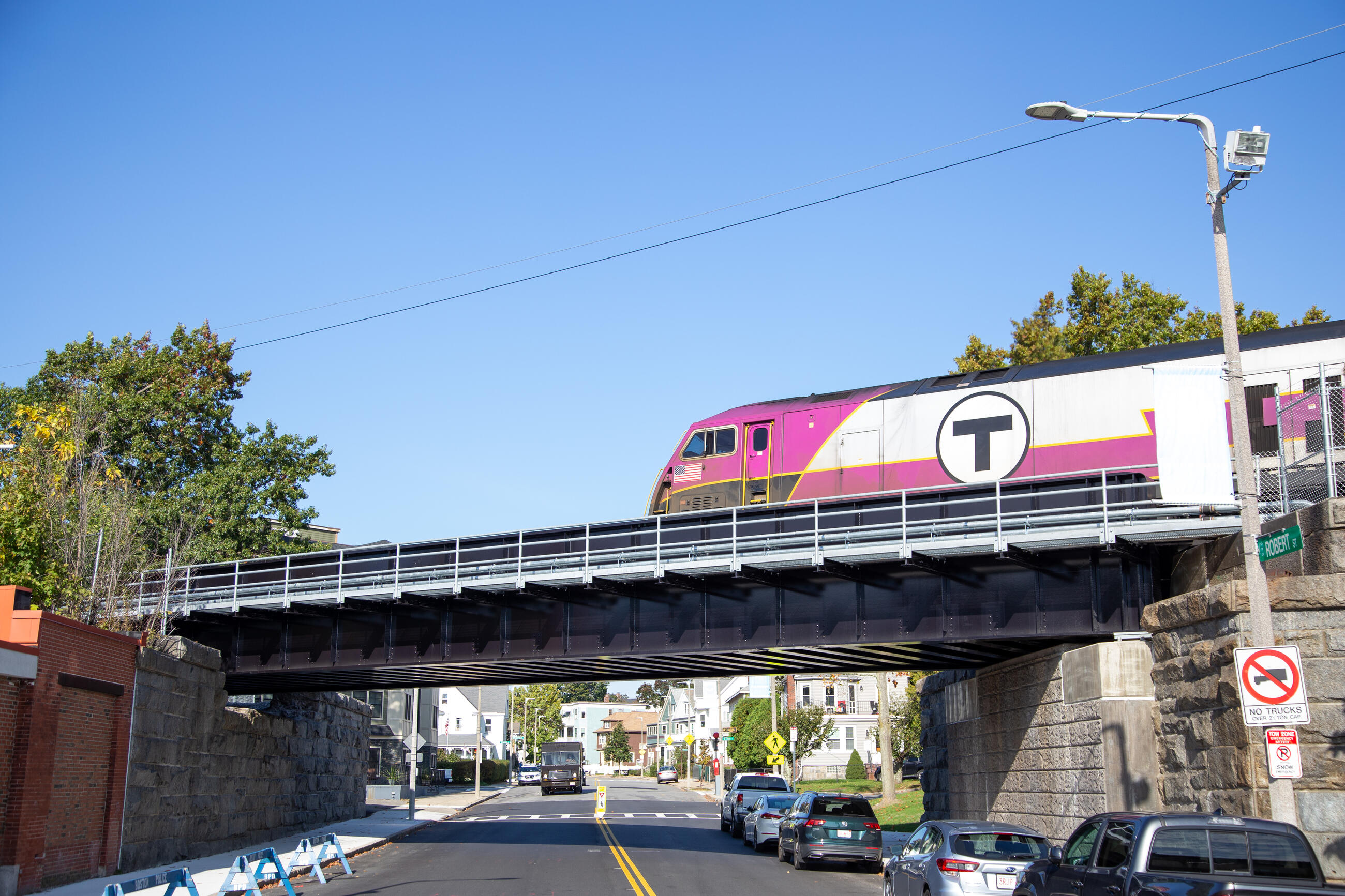 A Needham Line Commuter Rail train crosses the new Robert Street Bridge on a sunny day. A line of cars is parked under the bridge on Robert Street.