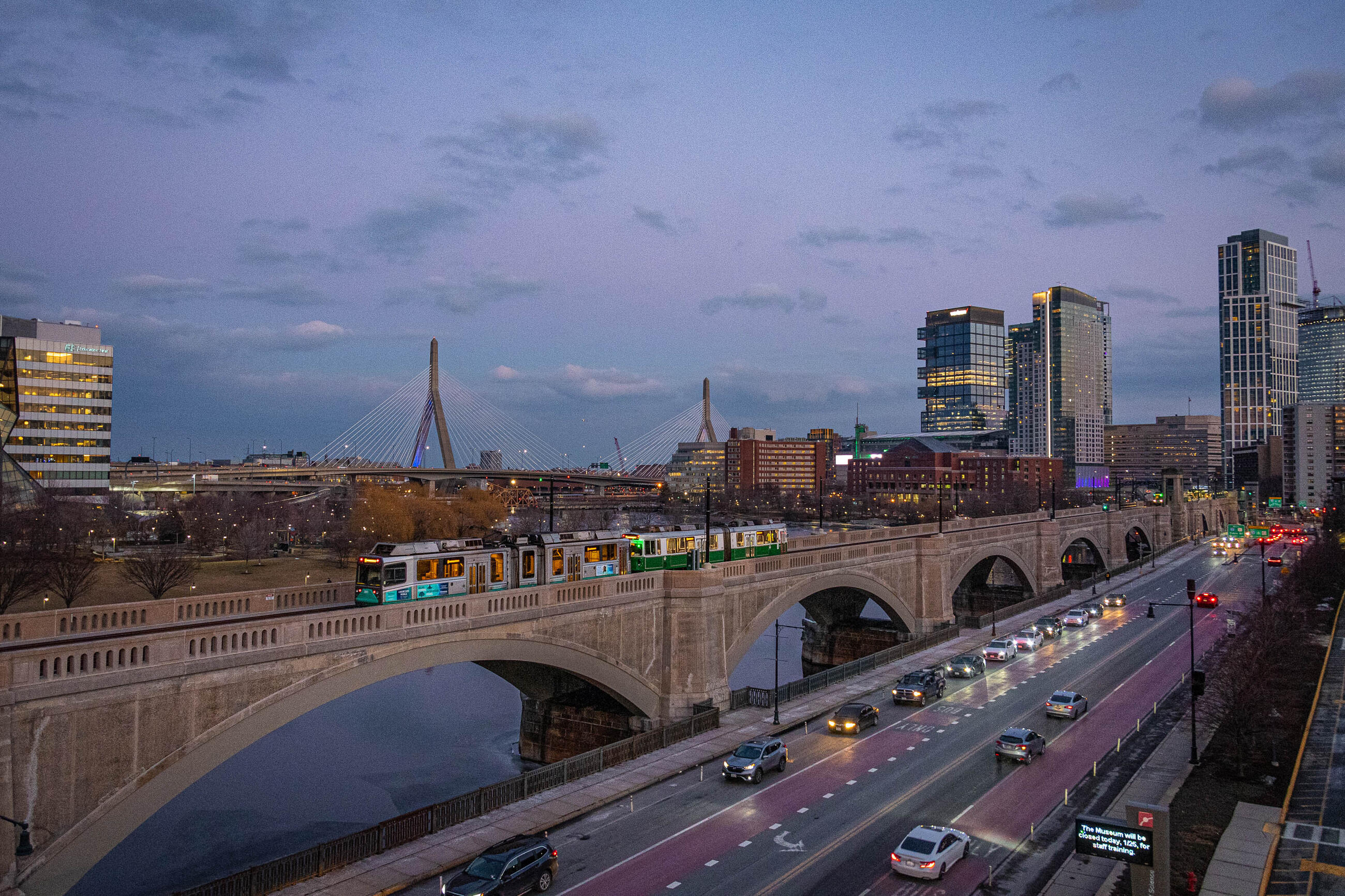 an empty green line train crossing the lechmere viaduct, with some boston skyline visible in the background, road traffic in the foreground, and a pretty pink and purple sunrise backdrop