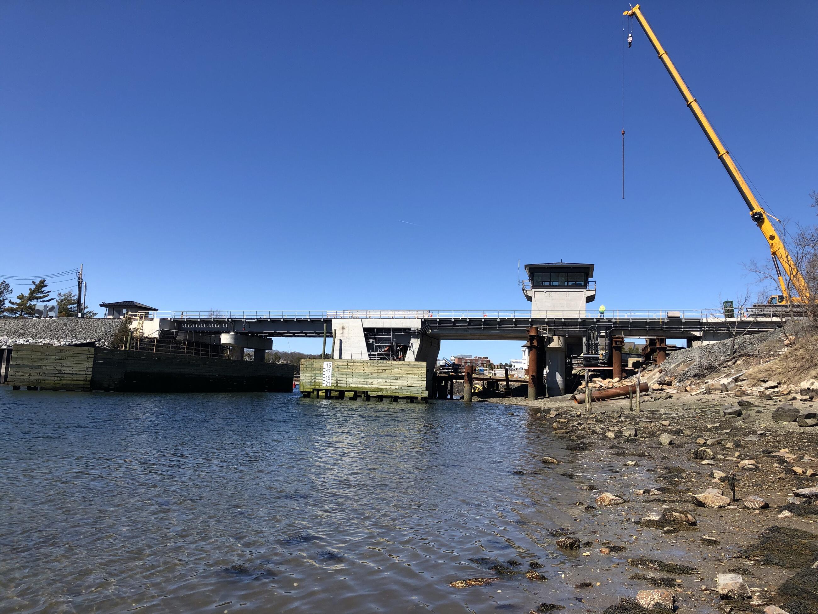 A wide view of the entire Gloucester Drawbridge under construction as seen in March 2022. A crane is at right suspending a long metal object in the air to the right of the control tower. At center bottom is the waterway over which the bridge spans. The bridge is in the middle distance under a dark blue sky. Both approaches to the bridge appear on either side of the photo.