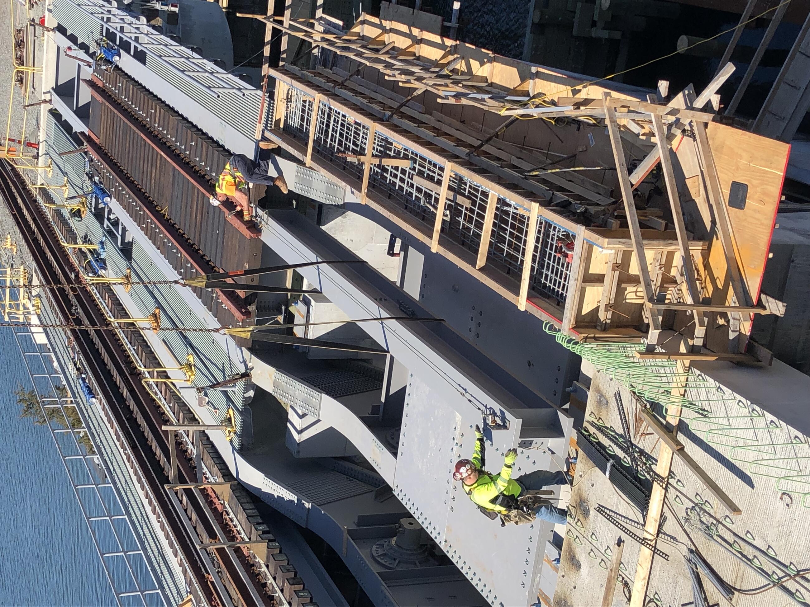 A long, elongated, box-shaped steel beam is suspended over the bascule counter weight on the bridge. Yellow-jacketed workers in hardhats hold each end of the beam and move it into place. The train tracks can be seen at upper right, the waterway at upper left. 