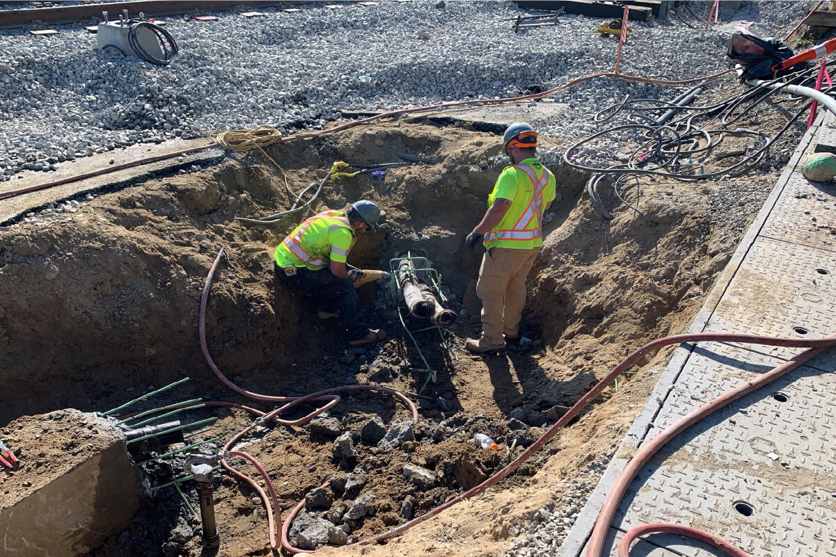 two crew members working on something in a shallow hole near the tracks