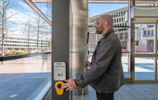 A blind man holding a white cane is seen in profile standing before a digital sign. His left hand is on a button labeled PRESS FOR AUDIO in standard writing and in braille. The rider stand in the lobby of the Government Center station; through the windows behind him the Government Center buildings are shown at right. 